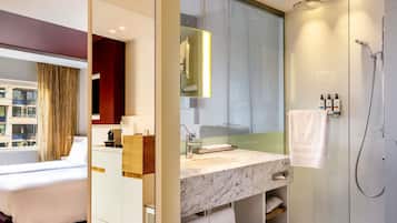 Una habitación de hotel moderna con una cama, un baño con ducha, un lavabo de mármol y un ventanal grande.