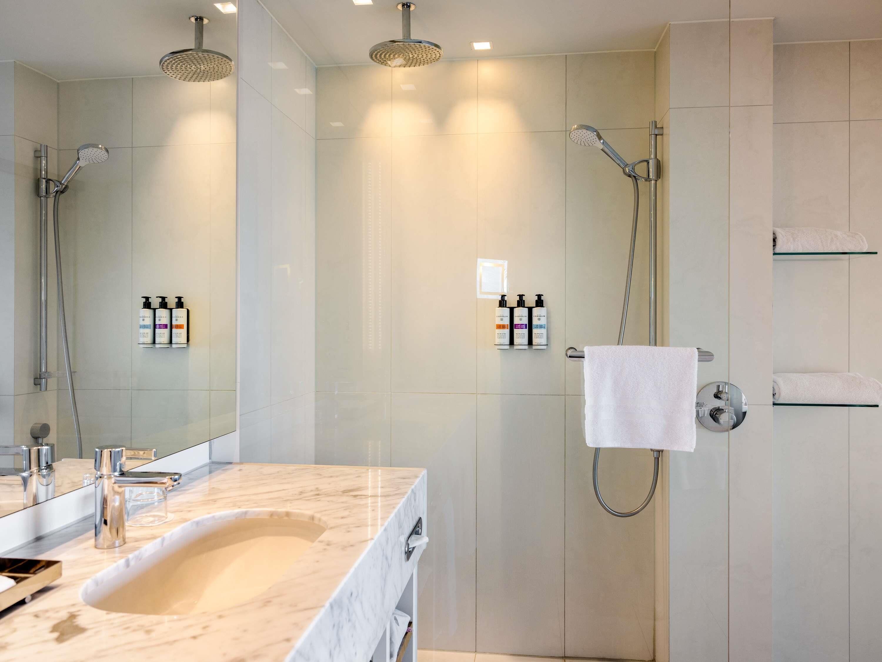 A modern bathroom with a marble sink, two overhead showers, and a glass shelf with toiletries.