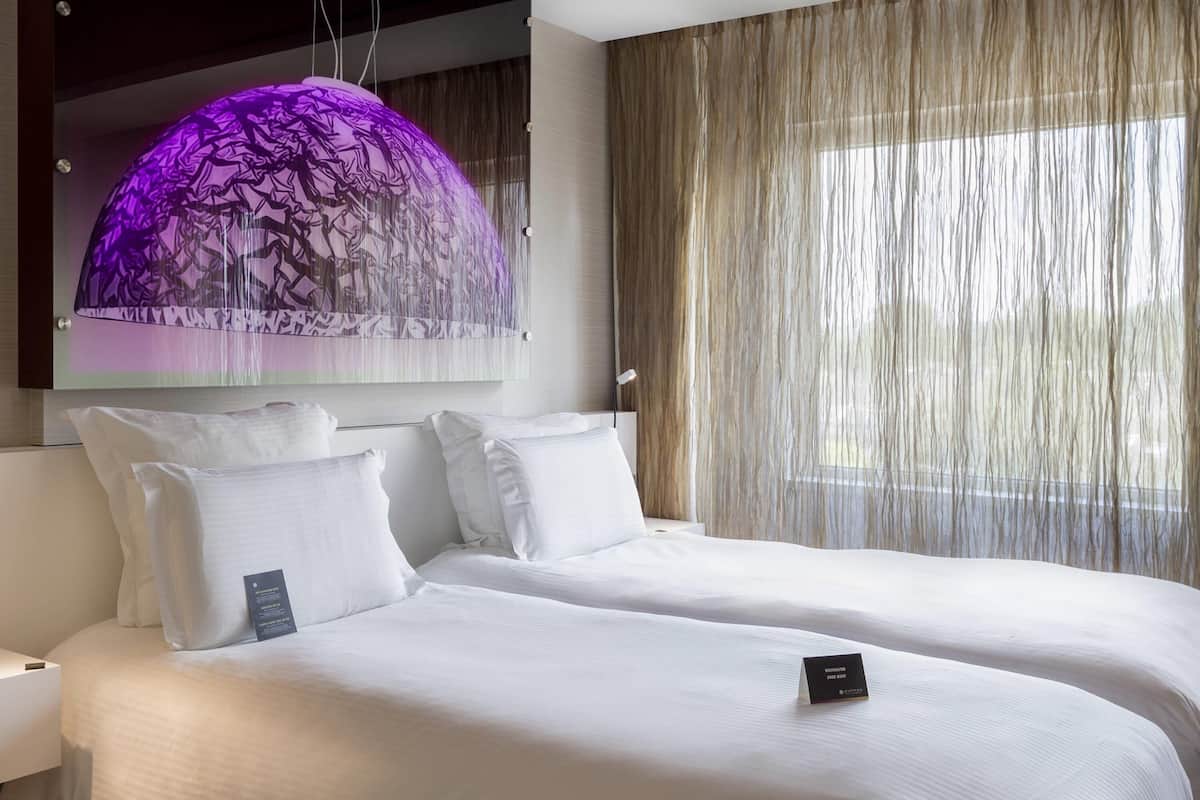 A hotel room with a large bed, a hanging light fixture, and a window with sheer curtains.