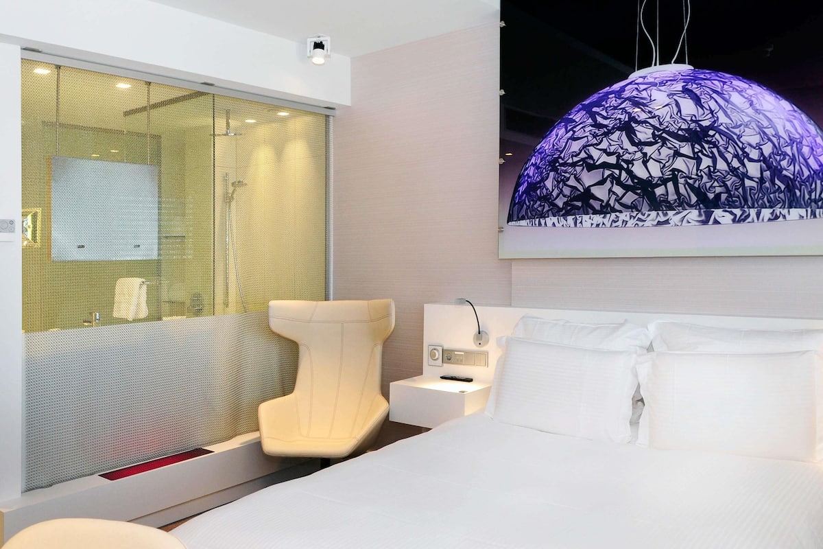 A modern hotel room with a large bed, a seating area with a chair, a bathroom with a glass partition, and a unique ceiling light fixture.