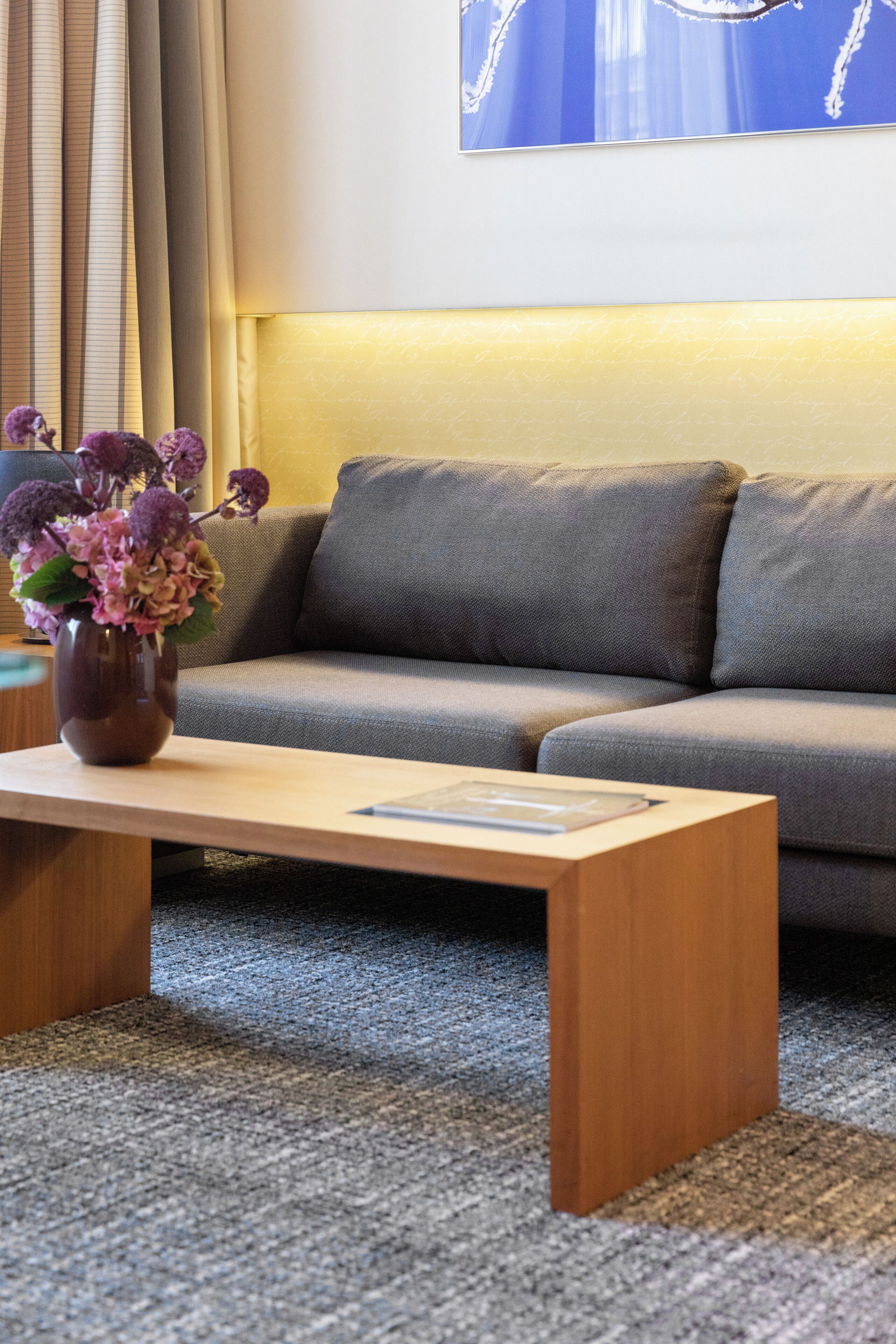 Een moderne woonkamer met een grijze bank, een houten salontafel en een vaas met paarse bloemen.
