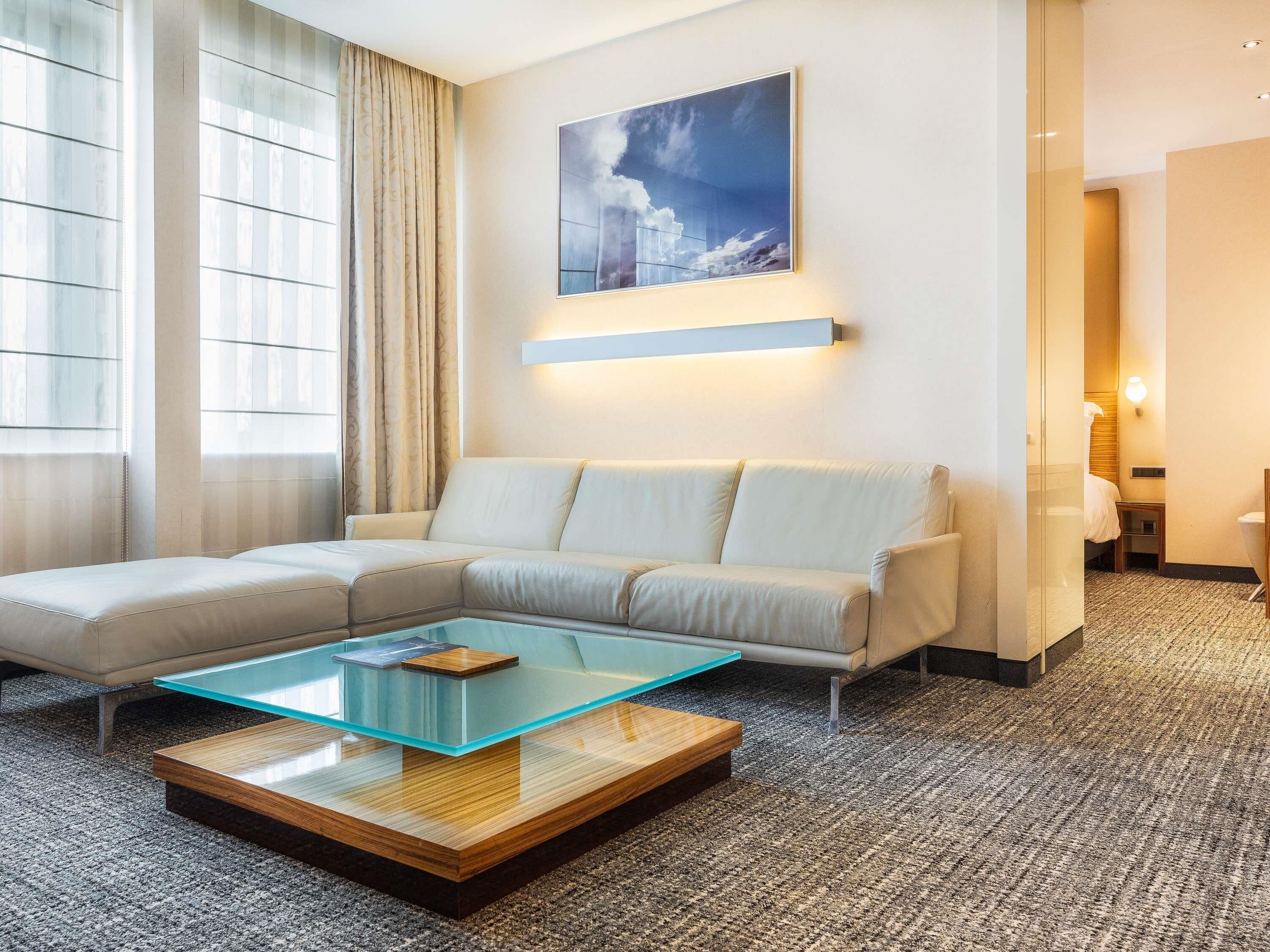 Une chambre d’hôtel moderne avec un canapé d’angle, une table basse en verre, un tableau au mur et une vue par la fenêtre.