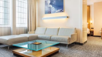 Une chambre d’hôtel moderne avec un canapé d’angle, une table basse en verre, un tableau au mur et une vue par la fenêtre.