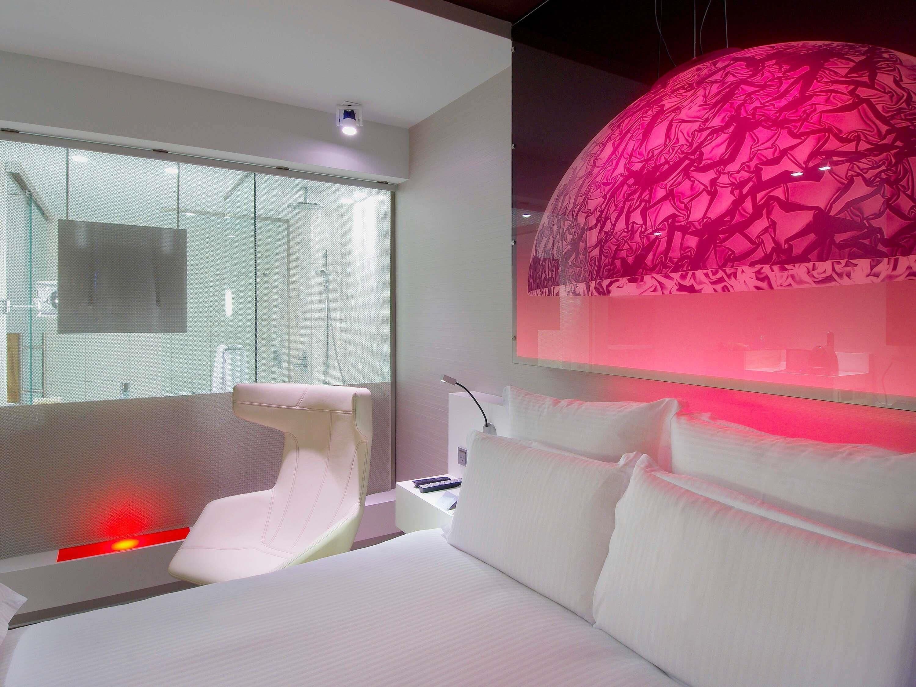 A modern hotel room with a large bed, a white chair, a glass shower enclosure, and a unique pink decorative element.