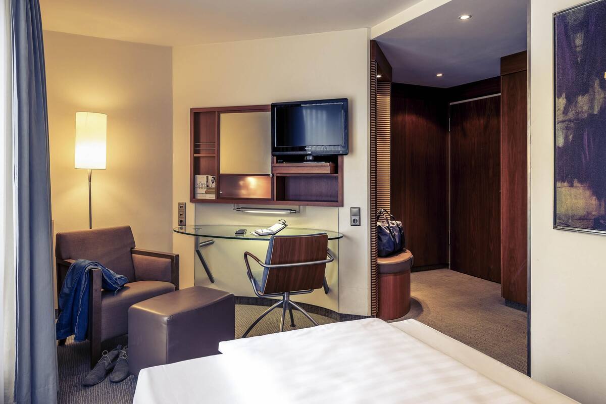 Ein Hotelzimmer mit Bett, Schreibtisch mit Computer, Stuhl, Fernseher, Lampe und einem Bild an der Wand.