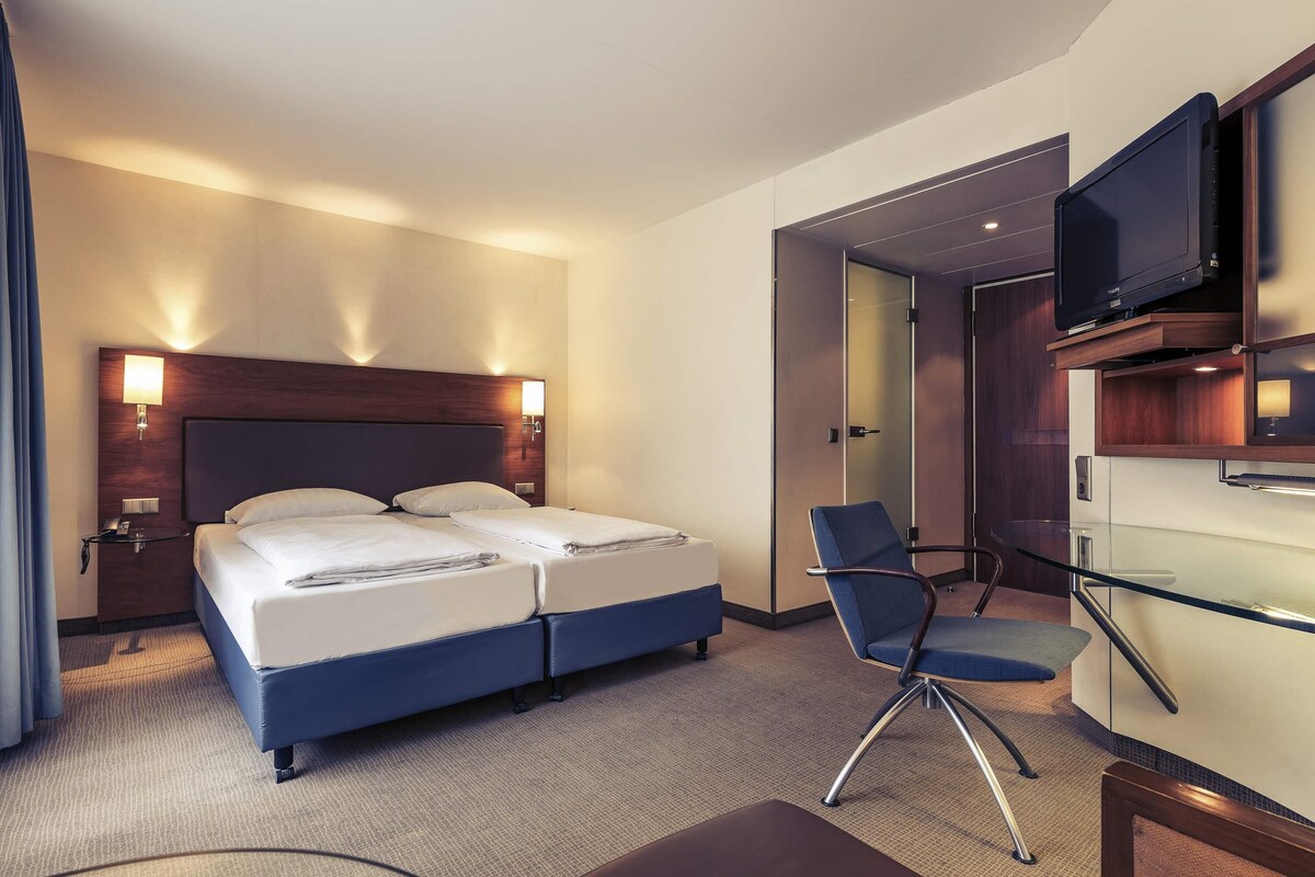 A hotel room with a bed, a desk, a chair, a TV, and a minibar.