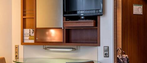 A hotel room with a glass desk, a television mounted on a wooden cabinet, and a wooden door with a window.