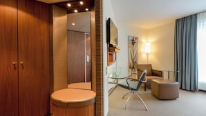 Ein Hotelzimmer mit Holzschrank, Glastisch, einem Sessel, einer Couch und einem an der Wand befestigten Fernseher.