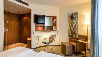 A hotel room with a bed, desk, chair, television, artwork, and a lamp.