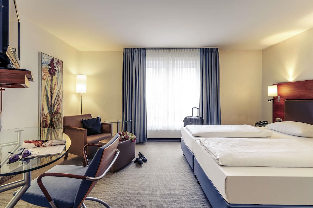 A hotel room with a bed, a desk with a chair, a small table, a lamp, a suitcase, and a painting on the wall.