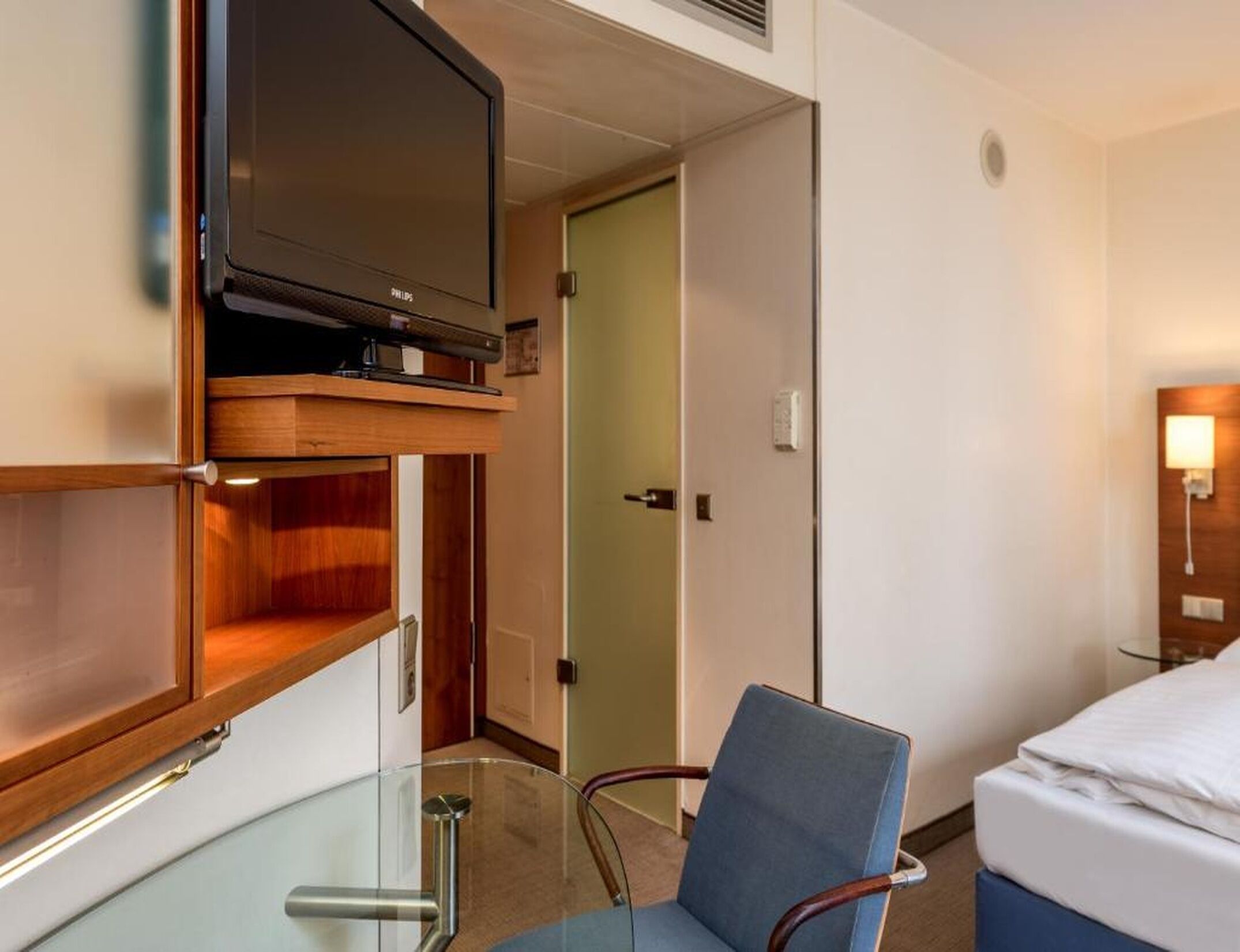 A hotel room with a bed, a television mounted on a wooden shelf, a glass table, and a chair.