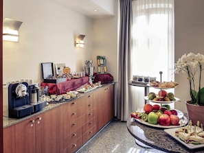 Habitación de hotel con desayuno tipo bufé, que incluye una cafetera, fruta, bollería y una orquídea ornamental.