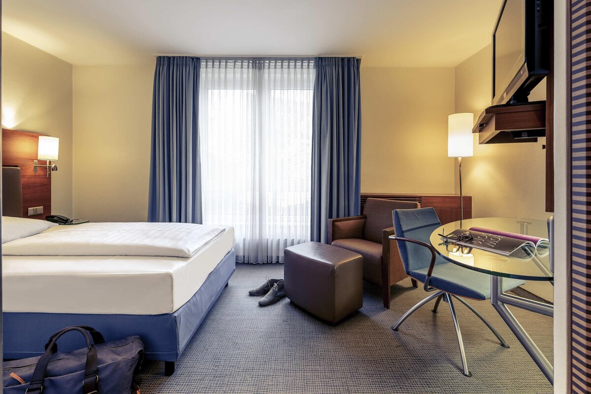 A hotel room with a bed, a desk with a chair, a small table, a lamp, and a window with curtains.