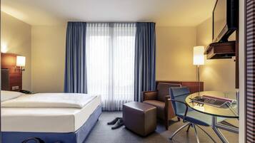 A hotel room with a bed, a desk with a chair, a small table, a lamp, and a window with curtains.