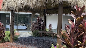 A thatched-roof structure in a garden setting with a gravel path, surrounded by various plants and a building with large windows in the background. - Vero Beach Inn & Suites I-95 (Vero Beach)