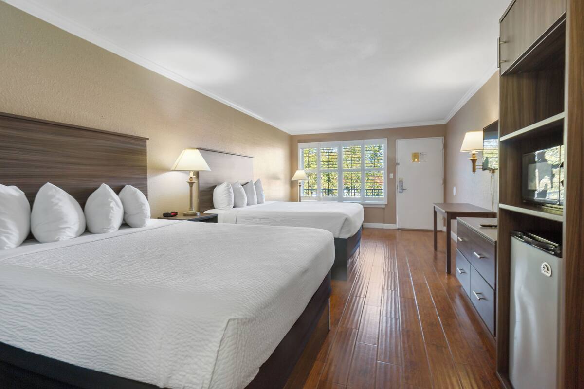 A hotel room with two beds, a wooden dresser, a desk, and a window with a view of trees.