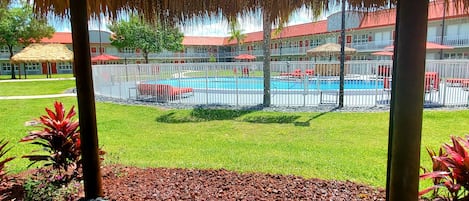 Un área de piscina con un pabellón de techo de paja, rodeada por una cerca blanca, y un edificio con tejados rojos al fondo.