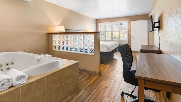 A modern hotel room with a large bathtub, a wooden dining table, and a bed with a view of greenery through the window. - Vero Beach Inn & Suites I-95 (Vero Beach)