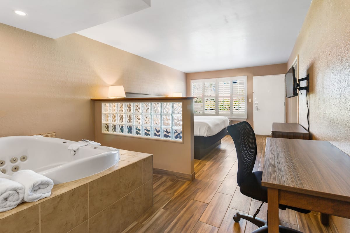 A modern hotel room with a large bathtub, a wooden dining table, and a bed with a view of greenery through the window.
