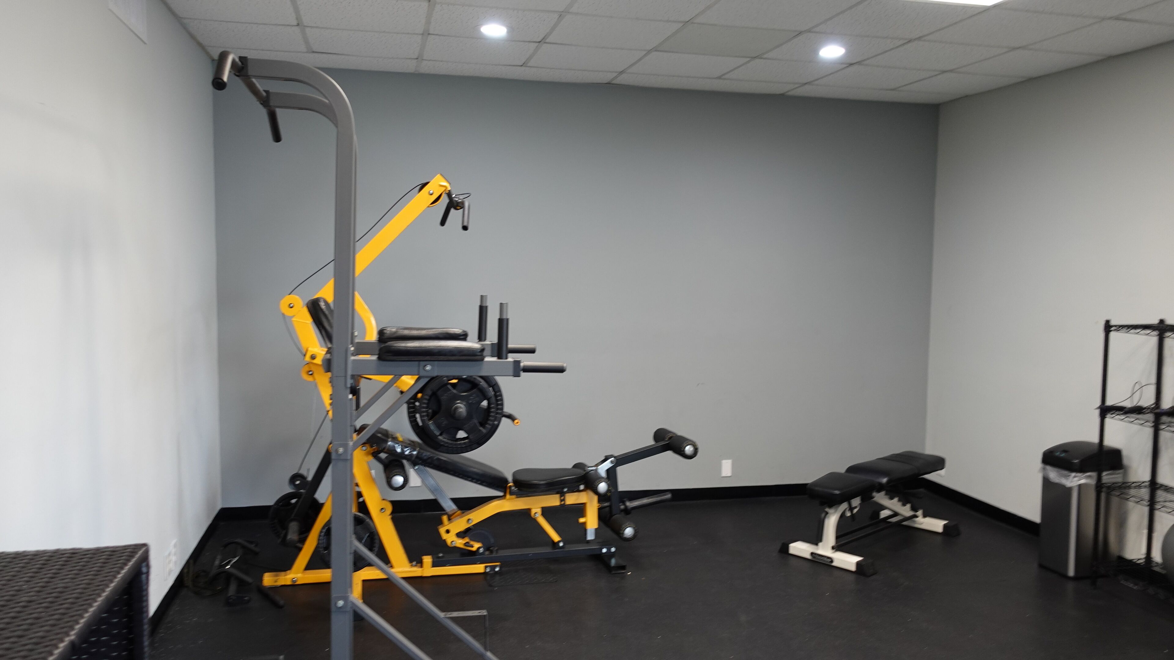 Un gimnasio con equipo de ejercicio, incluyendo una banca, máquina de pesas y un estante.