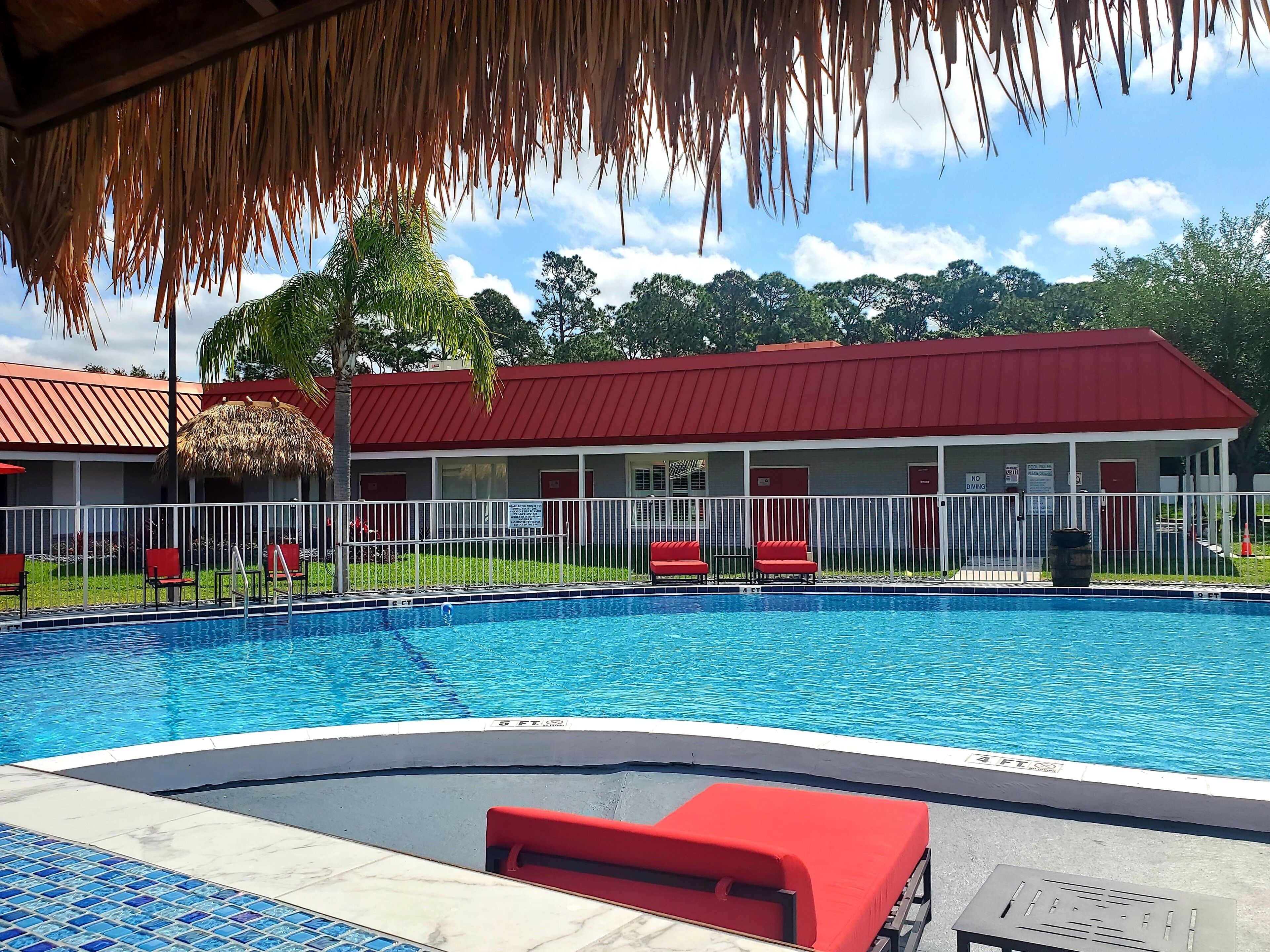 Une piscine avec un fauteuil de plage rouge, une structure à toit de chaume et, au fond, un bâtiment avec un toit rouge.