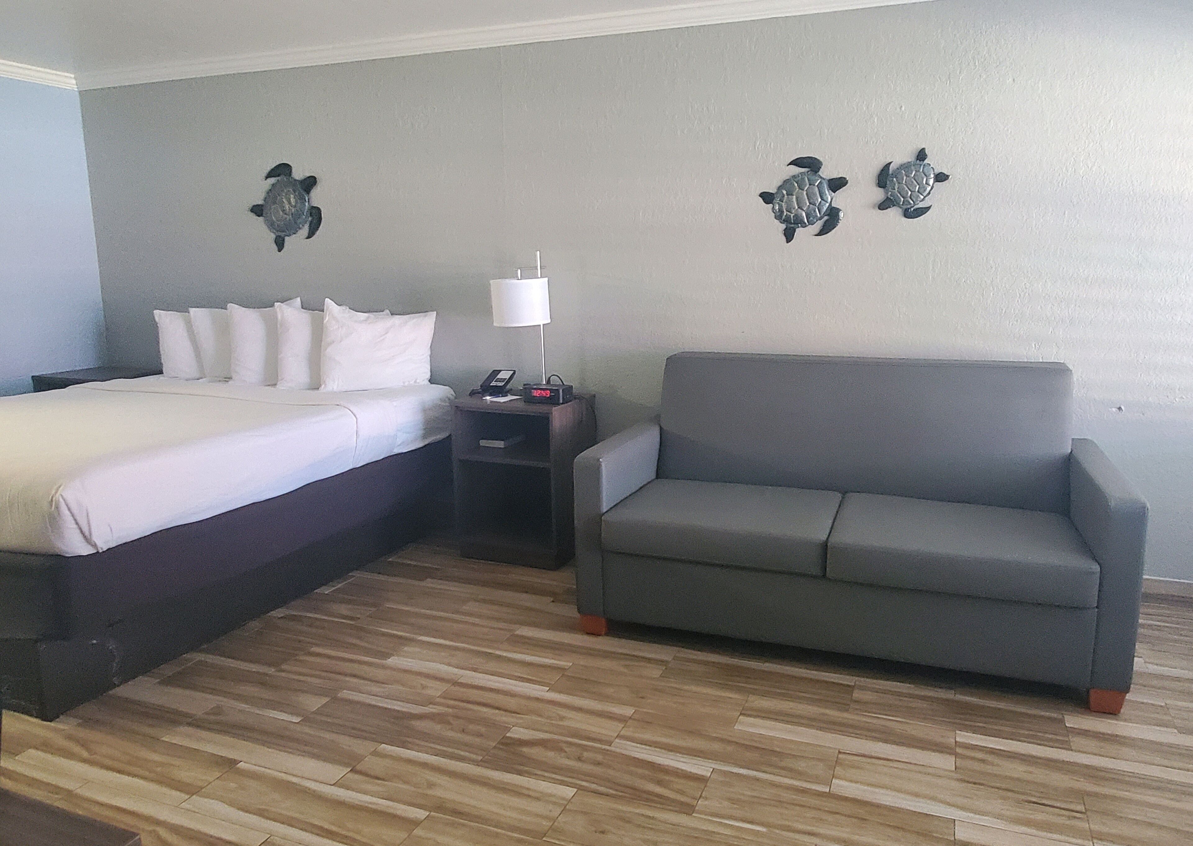 A hotel room with a bed, a grey sofa, a nightstand with a lamp, and two wall decorations.