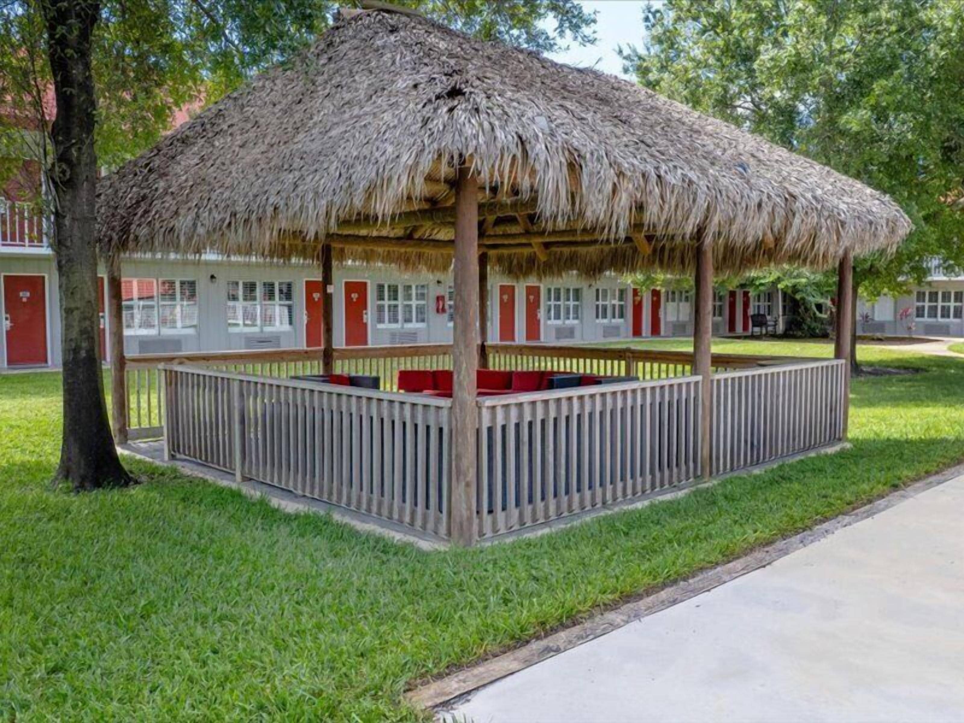 A thatched-roof gazebo with a wooden railing, surrounded by a lawn and a building with red doors in the background.