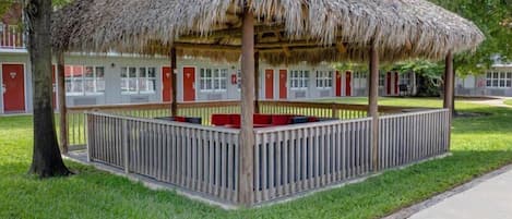 A thatched-roof gazebo with a wooden railing, surrounded by a lawn and a building with red doors in the background.