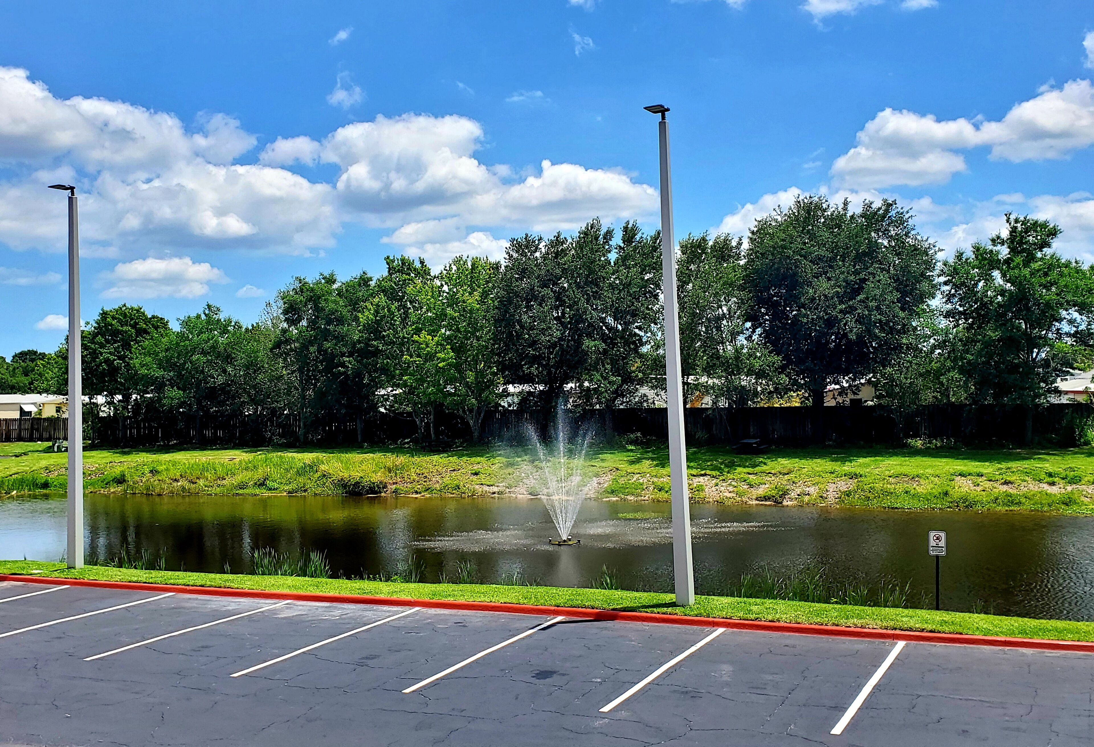 A parking lot with a pond and fountain, surrounded by trees and a clear sky.