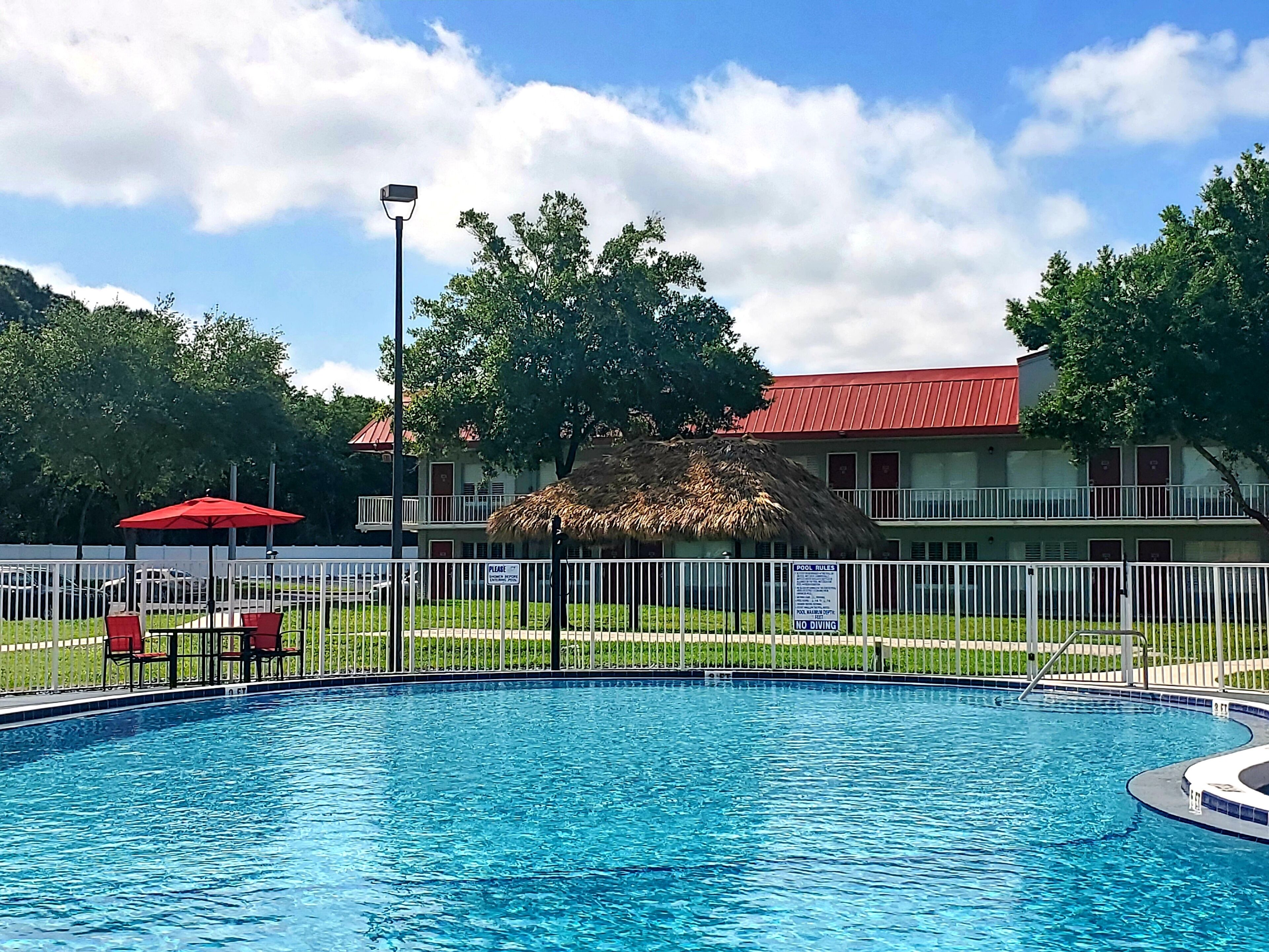 Una piscina con una cabaña de techo de paja, una sombrilla roja y una valla blanca.