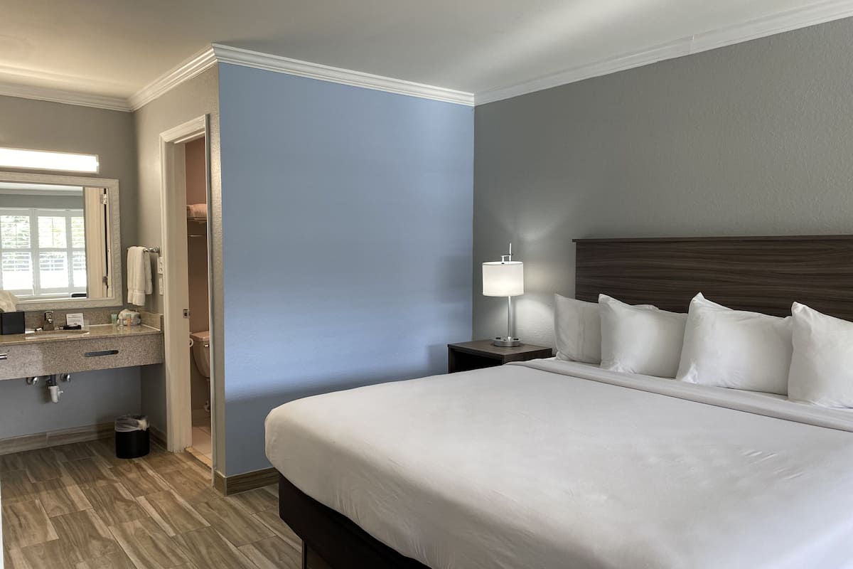 A hotel room with a large bed, a nightstand with a lamp, a bathroom with a sink and mirror, and a wooden floor.