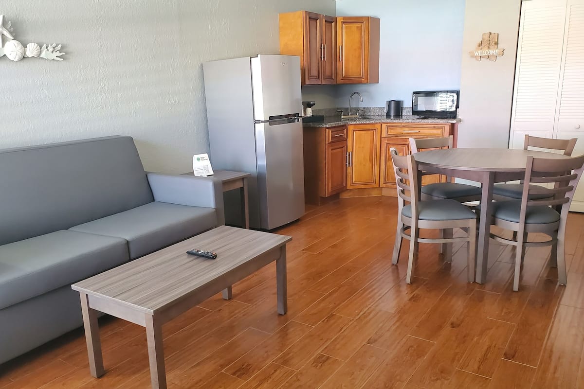 A modern living area with a grey sofa, a wooden table, and a kitchen with wooden cabinets and a refrigerator.