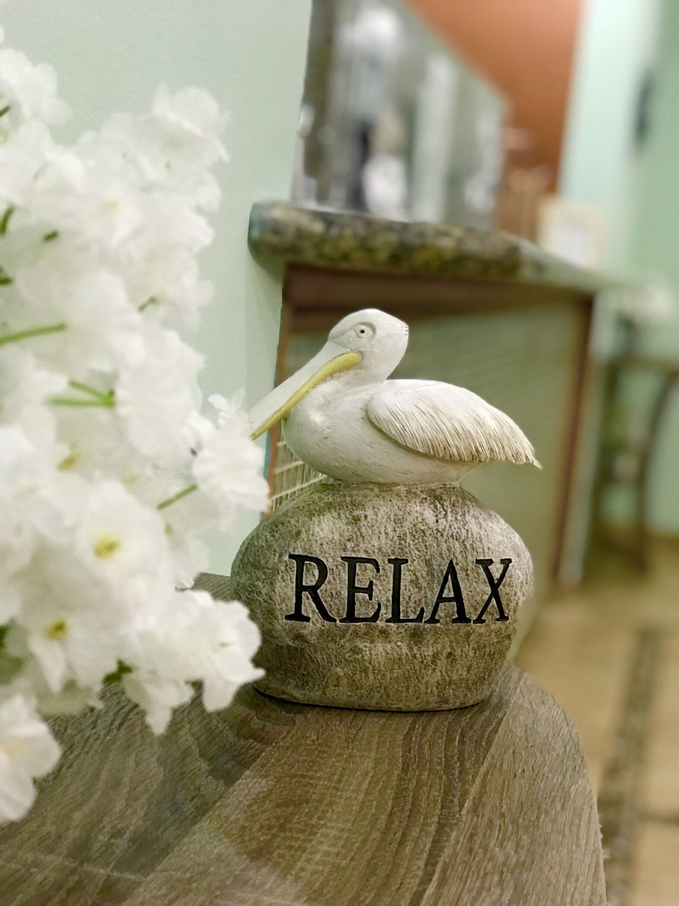 Kivestä veistetty pelikaanipatsas, joka on sijoitettu kalliolle. Patsaan kylkeen on kaiverrettu sana ”RELAX”. Patsas on asetettu puupinnalle, ja sen lähellä on valkoisia kukkia.