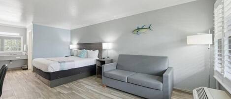 A hotel room with a bed, a gray sofa, a nightstand, a lamp, a wall decoration, and a window.