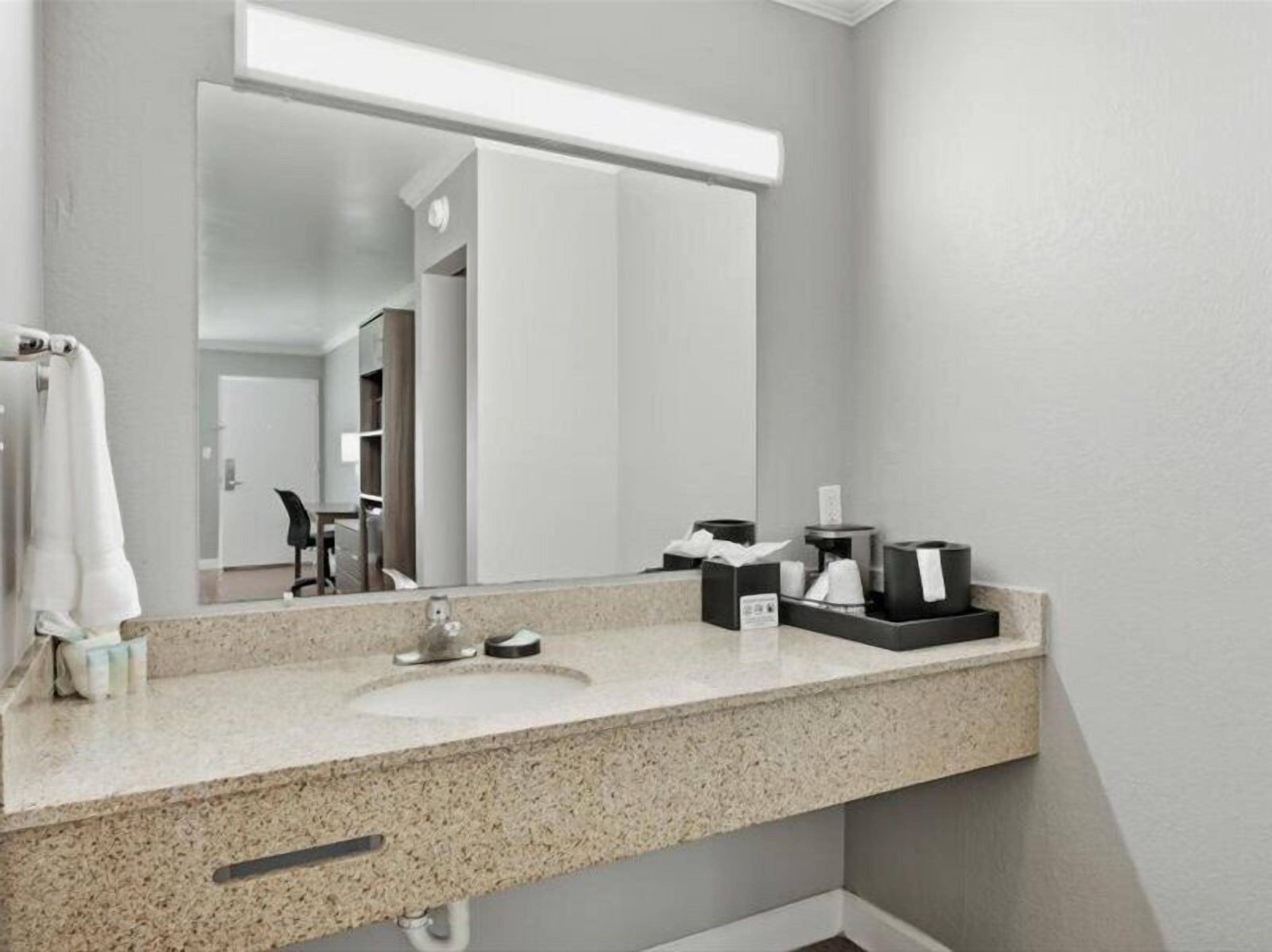 A hotel bathroom with a granite countertop, a sink, a mirror, and a towel rack.