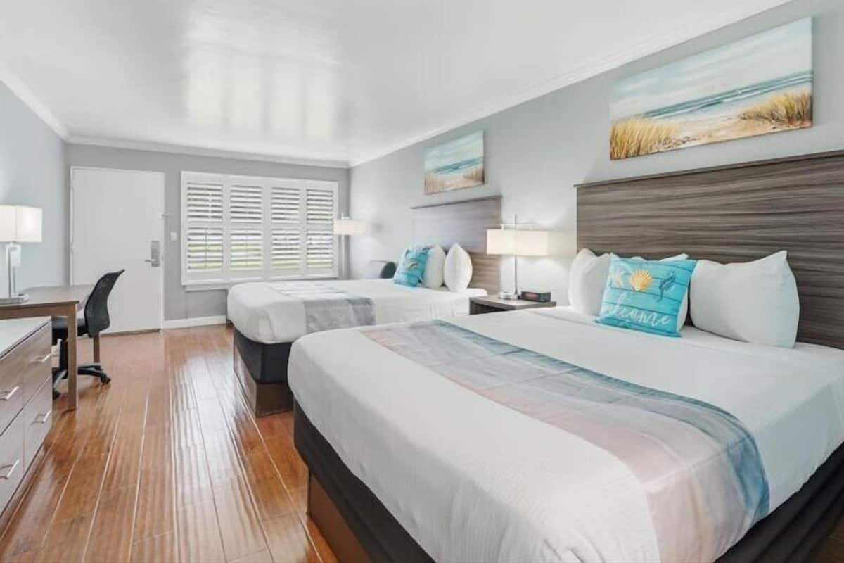 A hotel room with two beds, a desk, and a chair. There are two windows with blinds, a painting on the wall, and a lamp on the nightstand.