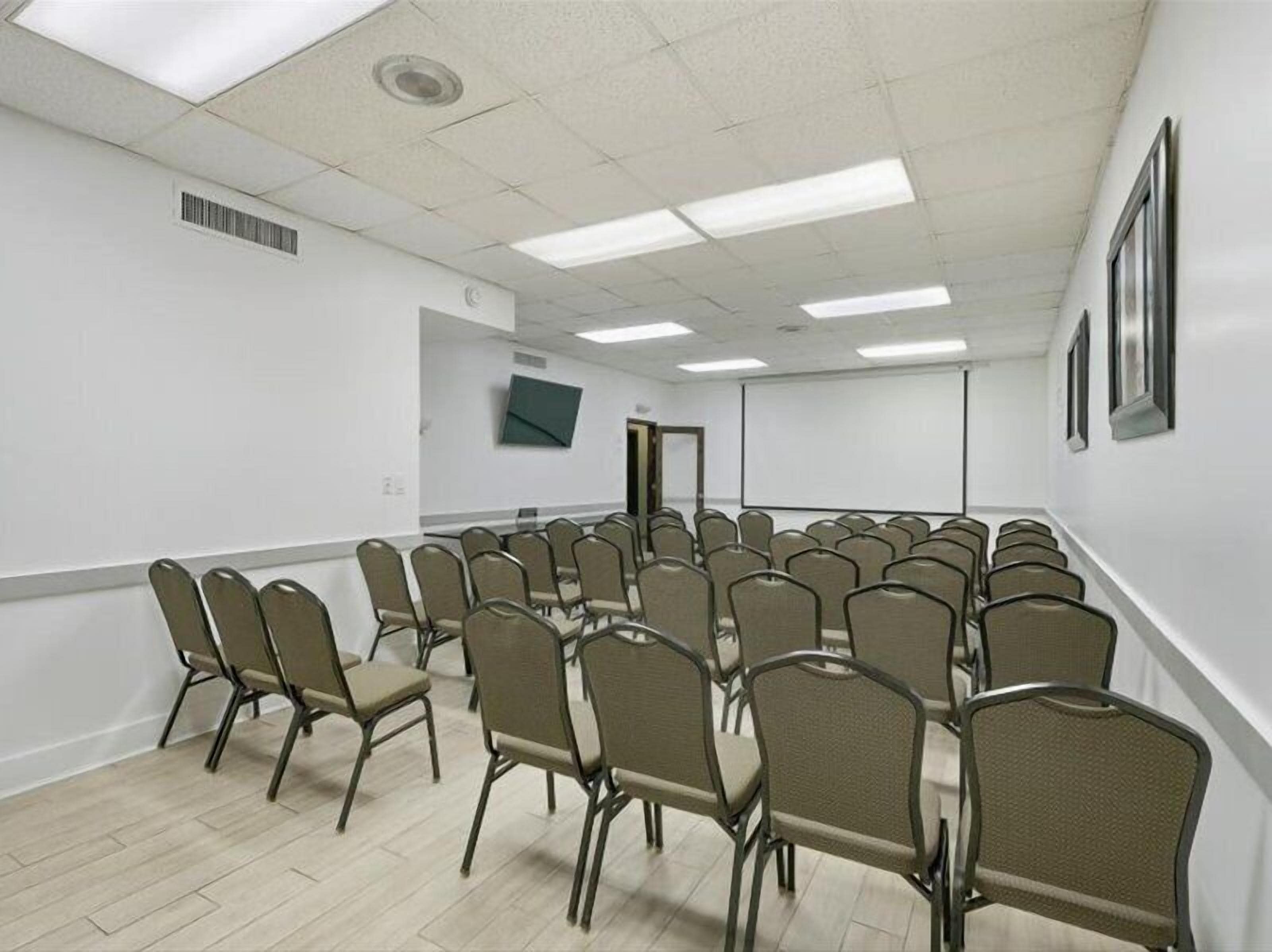 Un salón de conferencias con filas de sillas, una pantalla de proyector y un televisor en la pared.