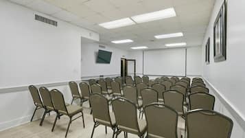 Un salón de conferencias con filas de sillas, una pantalla de proyector y un televisor en la pared.