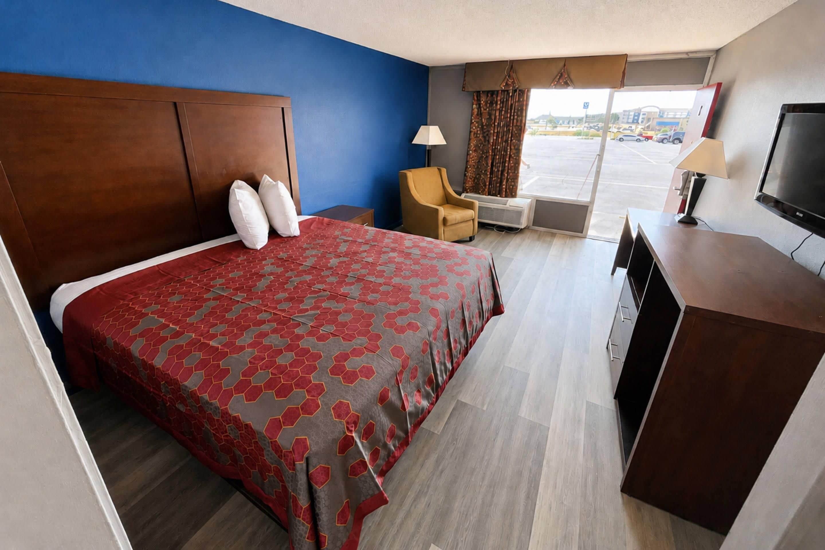 A hotel room with a large bed, a desk, a chair, a TV, and a window with a view of the parking lot.