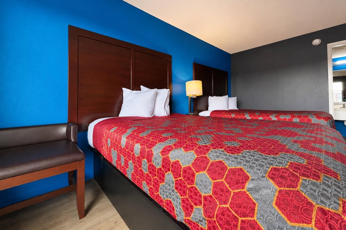 A hotel room with a large bed, a wooden headboard, a bedside table with a lamp, a bench, and a mirror on the wall.