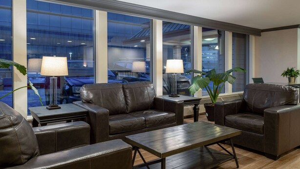 A modern hotel lobby with leather sofas, a wooden coffee table, and two lamps. Large windows offer a view of a parking garage. - Ramada by Wyndham Juneau (Juneau)