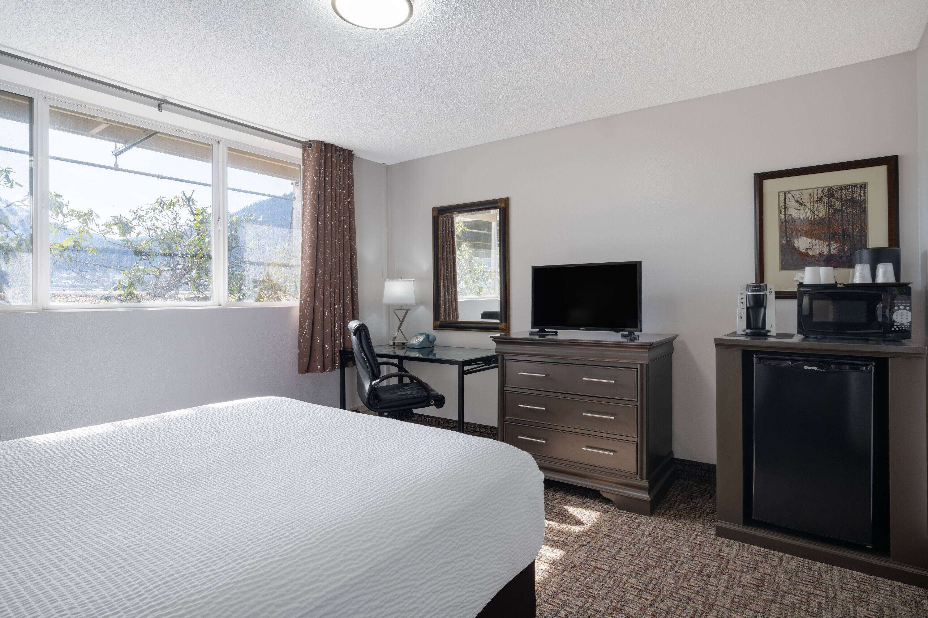 A hotel room with a large bed, a desk with a chair, a television, a coffee maker, and a refrigerator.