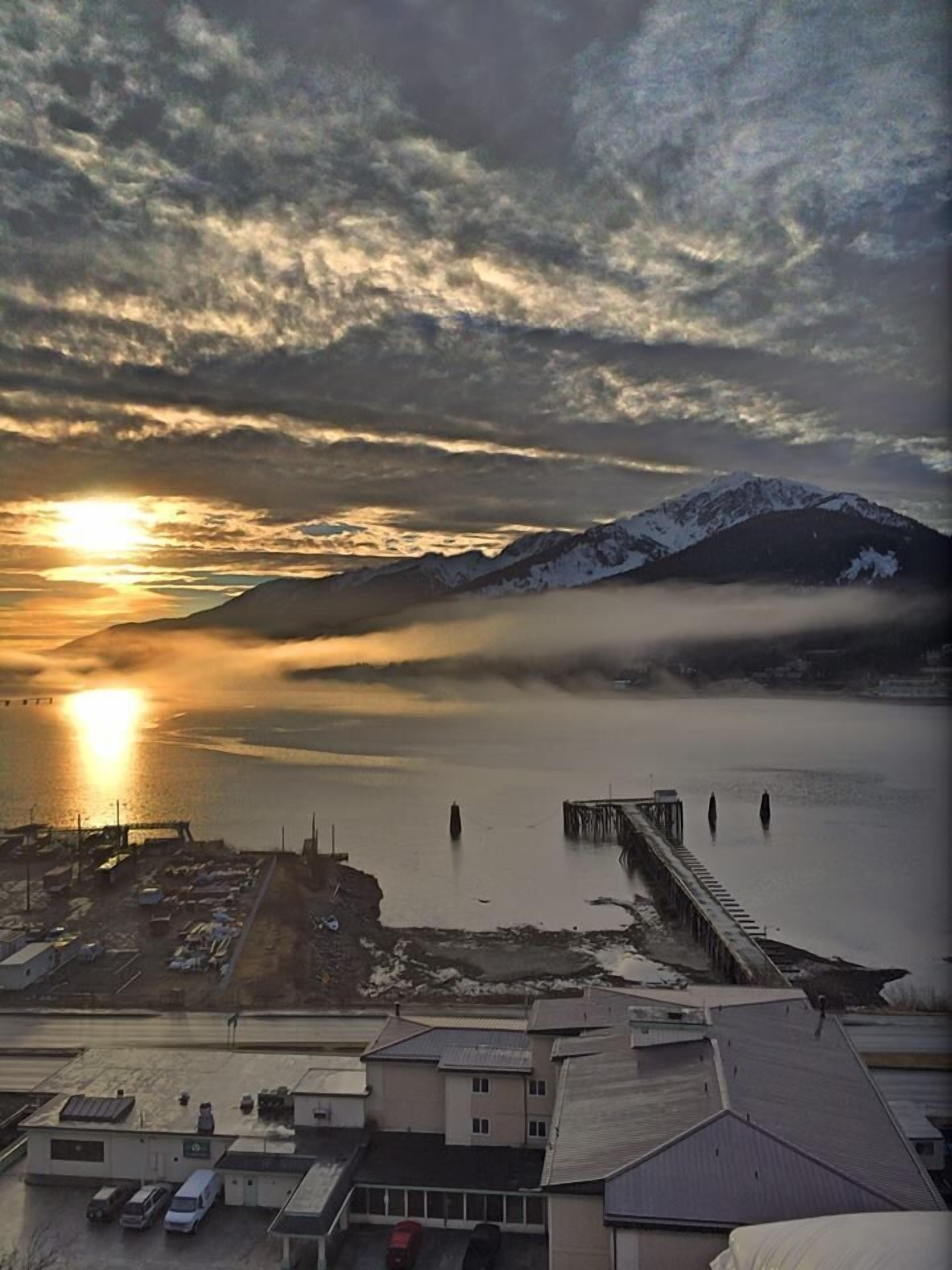Une ville côtière dotée d’un quai s’étendant dans des eaux calmes, entourée de montagnes et surplombée d’un ciel nuageux.