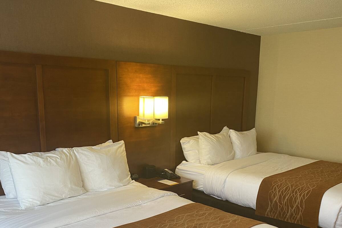 A hotel room with two beds, each with white bedding and brown decorative pillows. There is a wall-mounted light fixture above the beds.
