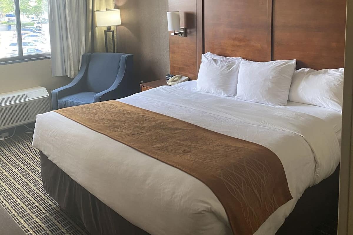 A hotel room with a large bed, a desk, a chair, a window with curtains, and a wall-mounted lamp.