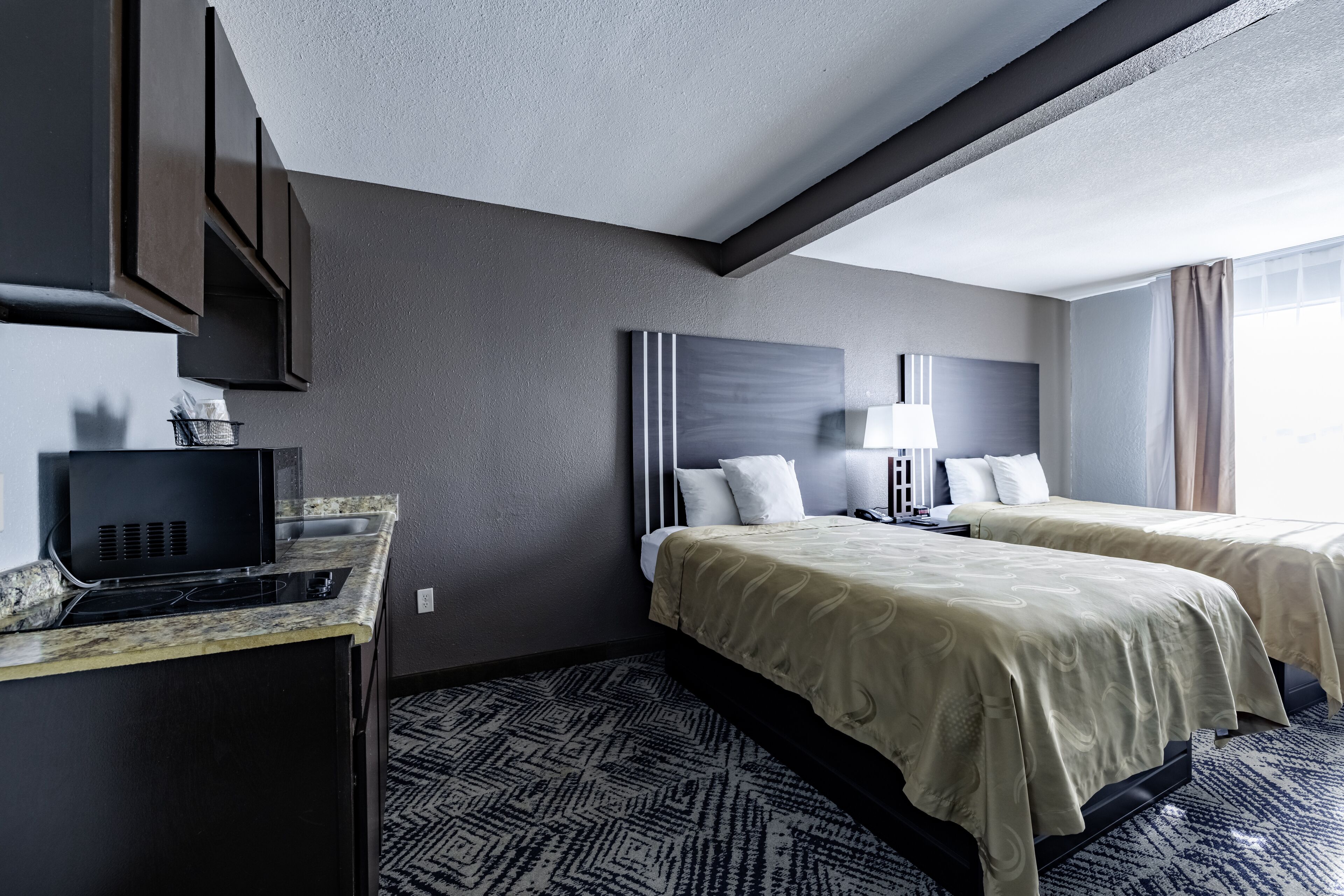 Habitación de hotel con kitchenette, dos camas, un ventanal grande y alfombra estampada.