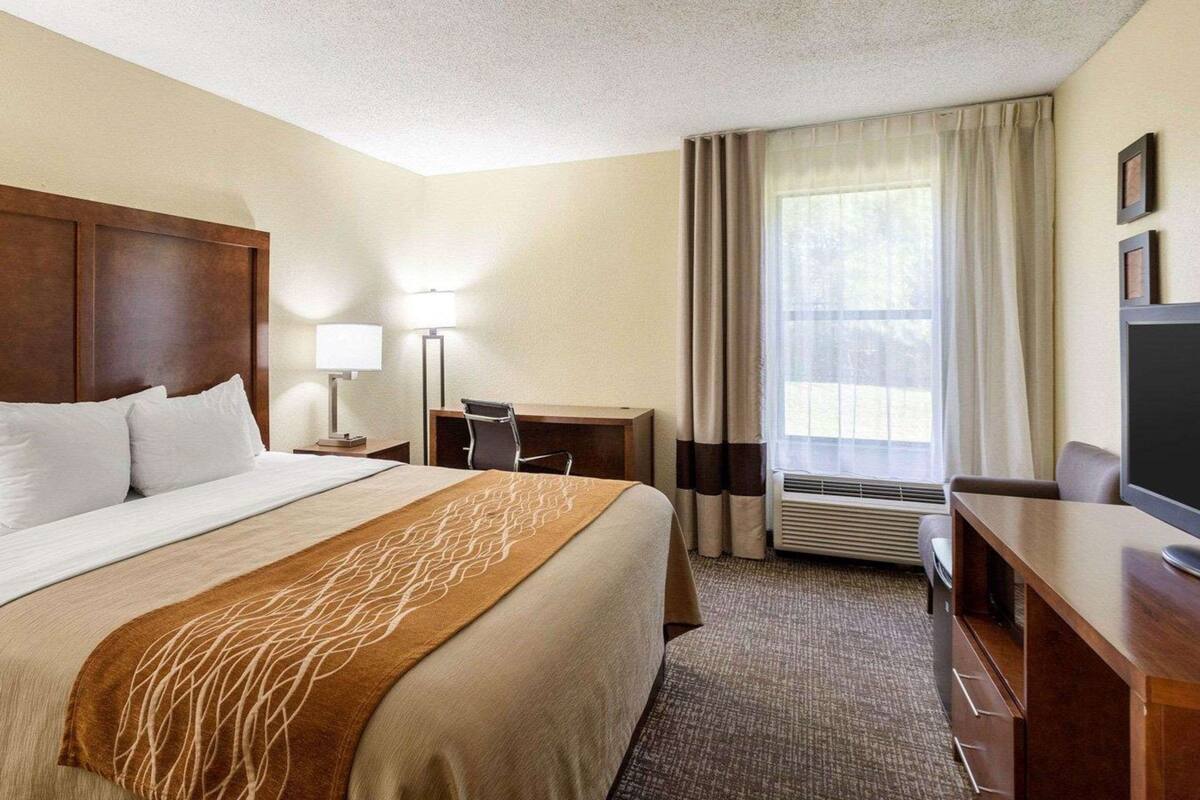 Ein Hotelzimmer mit Bett, Schreibtisch, Stuhl, Fernseher und einem Fenster mit Vorhängen.