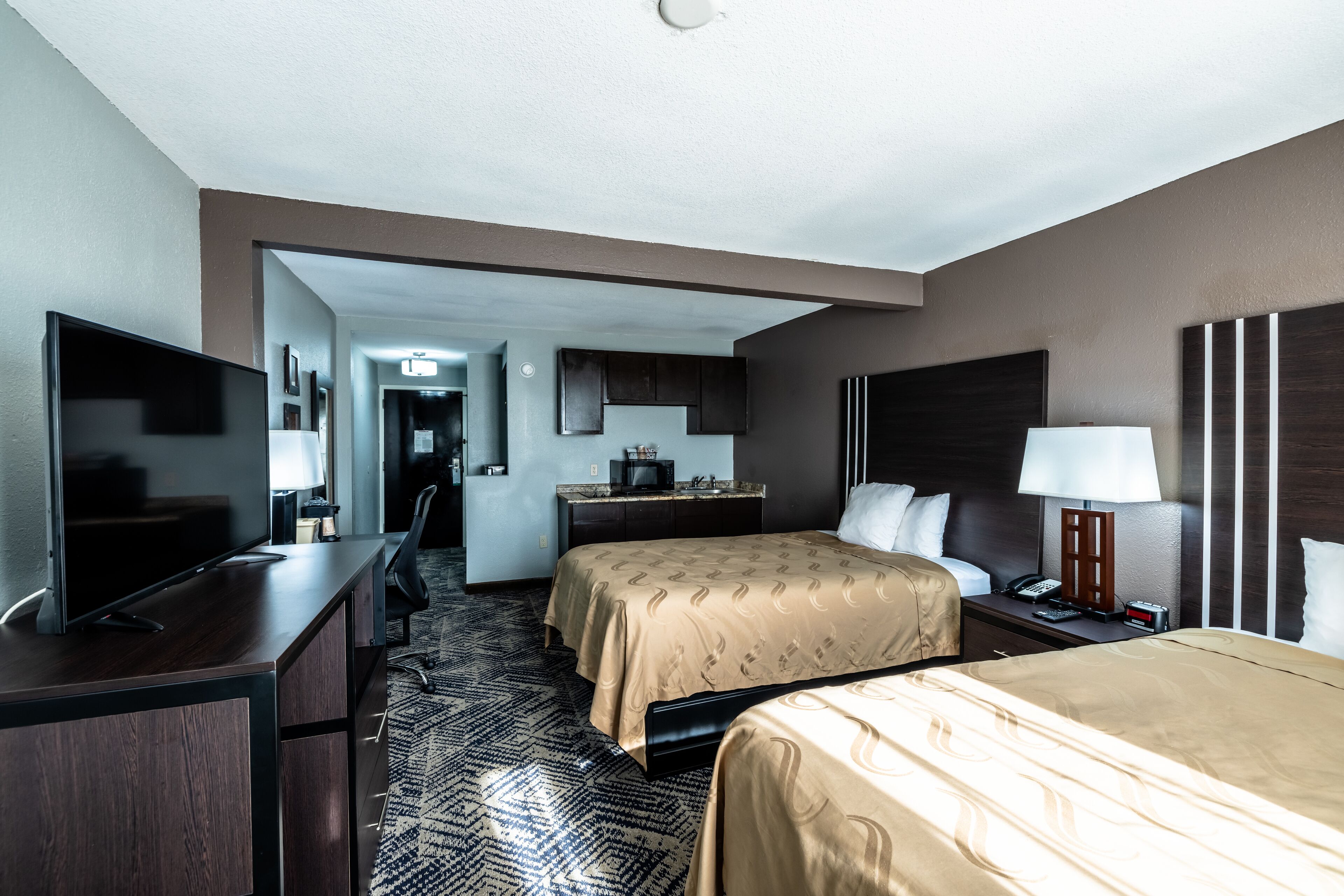 A hotel room with a bed, a desk with a TV, a bedside table with a lamp, and a bathroom visible through an open door.