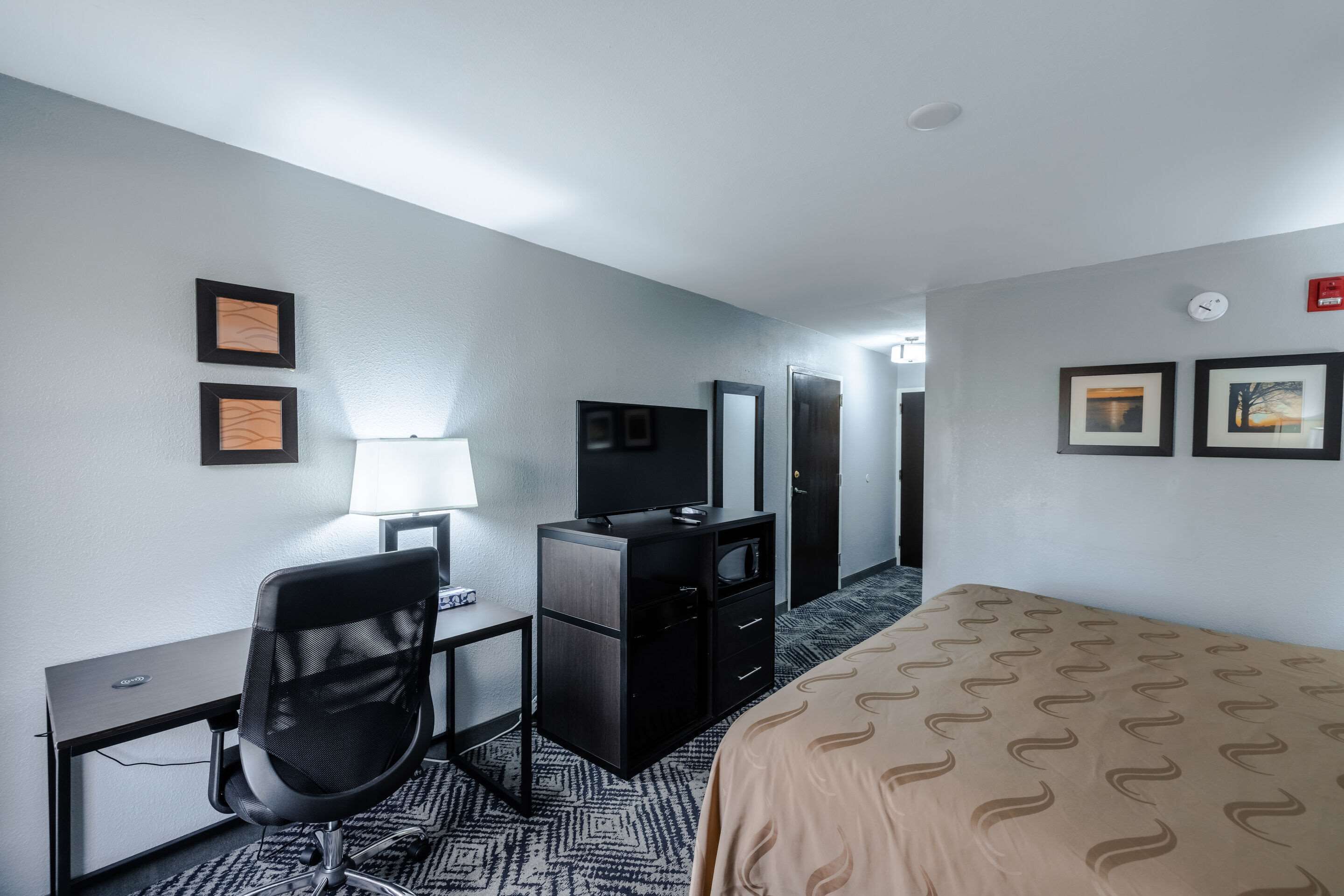 A hotel room with a bed, a desk with a chair, a dresser, a television, and a lamp.
