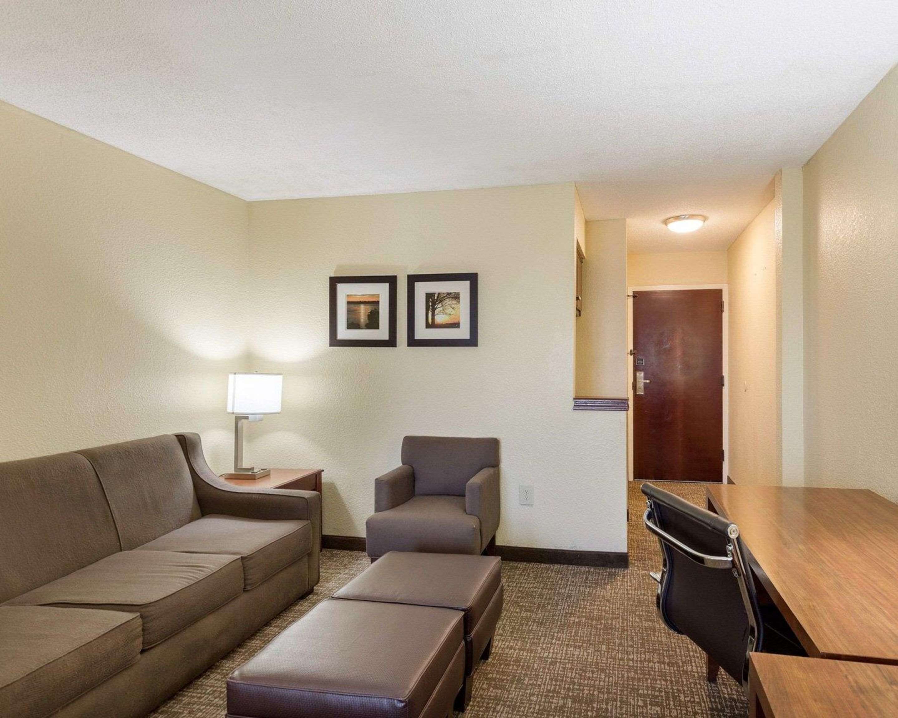 Ein Hotelzimmer mit einer Couch, einem Sessel, einem Hocker und einem kleinen Tisch. An der Wand hängen zwei gerahmte Bilder, eine Lampe und eine Tür zu einem weiteren Raum.